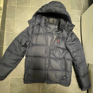 Ralph Lauren dunjacka  - Snygg mörkblå dunjacka från Polo Ralph Lauren med röd broderad logga på bröstet. Jackan har huva, flera dragkedjor och knappar vid kragen. Puffad design och praktiska fickor med dragkedja. Perfekt för kalla dagar. Jackan är använd men fortfarande gott skick! Säljer den nu då jag är sugen på en ny vinterjacka!