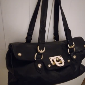 DKNY / Donna Karan New York svart handväska, guldiga detaljer, läder, fint skick - DKNY / Donna Karan New York svart handväska i äkta skinn/läder med snygga guldfärgade detaljer+tidningsmönstrat foder. Har totalt 4 fack inuti väskan, 2 små fickor och 2 lite rymligare fack med dragkedjor för tex plånbok, mobil, nycklar etc. 35x25x15 cm.