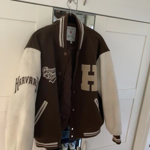 Varsity Jacka harvard - En brun varsity jacka med snyggt Harvard tema. Den är beige och brun o gräddvit. Kan lätt matchas med mycket snygga outfits och den är dessutom väldigt varm och skön. Perfekt höst/vårjacka  Mycket bra skick. Inga fel eller skador