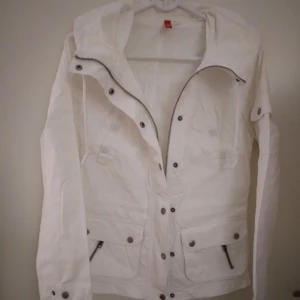 Vit vårjacka / sommarjacka / utilityjacka. Strlk: XS. Liten defekt. - Vit fin vårjacka / sommarjacka.  Stängs med dragkedja  och knappar, samt har dragsko i midjan + flera fickor. Strlk: XS.  Material: Bomull. Fint skick i övrigt MEN har en liten fläck bak på kragen. Går förhoppningsvis bort med klorin. FRAKT TILLKOMMER.
