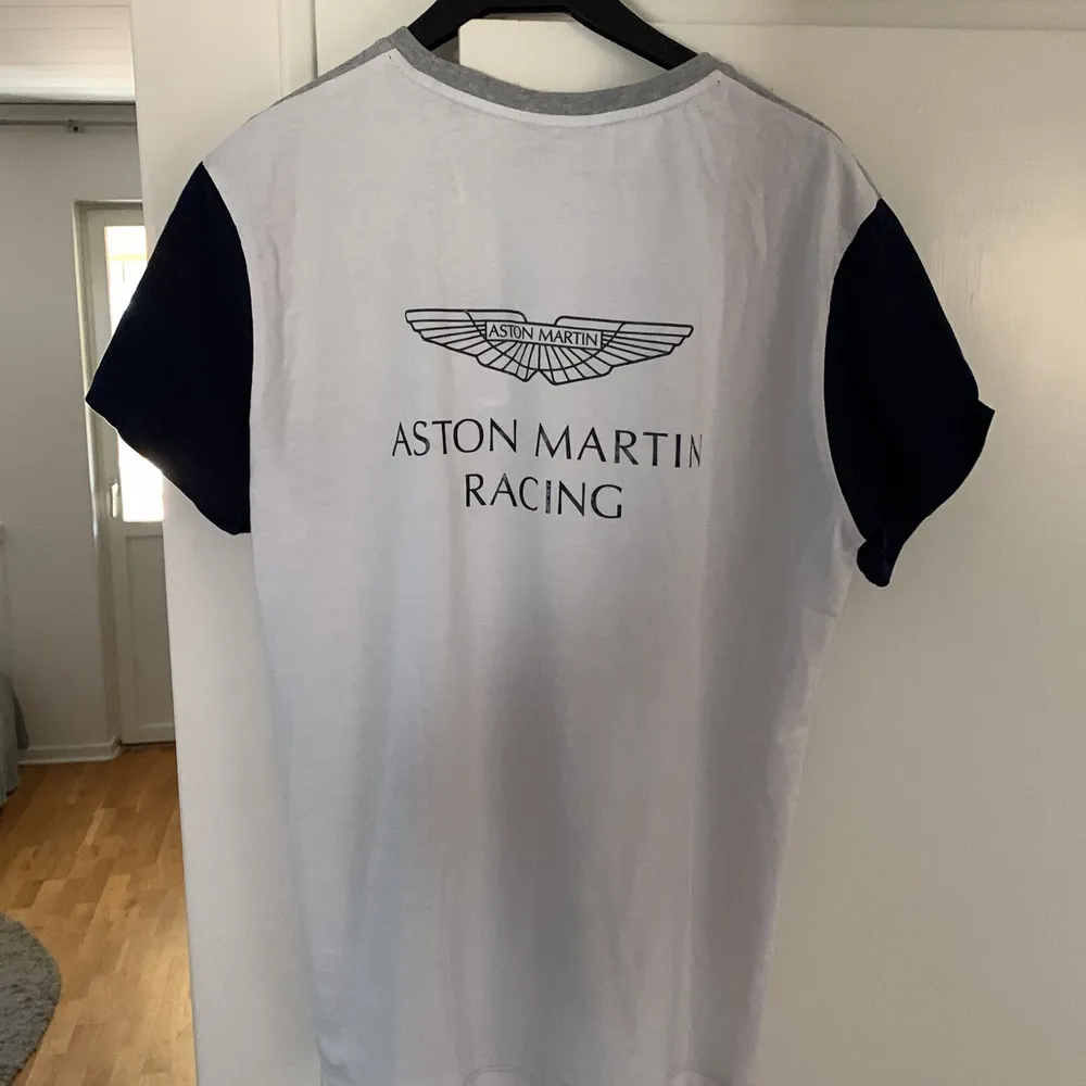 Fin och fräsch t-shirt från Aston Martin Formel 1 merch. Från märket Hackett som är känd för högkvalitativa kläder. Vit på baksidan och grå på framsidan, storlek M, något mindre än en vanlig medium. T-shirts.