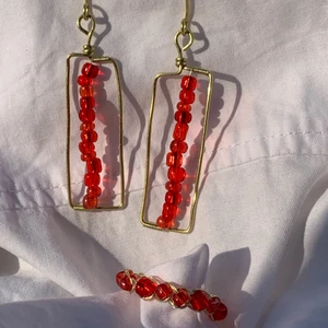 Smycken - Ett handgjort kit med örhängen och en ring. Gjorda med röda pärlor och guldpläterad koppartråd.  Nickel-, kadium-, och blyfria.  5,5x1,5 cm Ringarna finns i: 17mm, 18mm, 19mm (Innerdiameter av ringen)