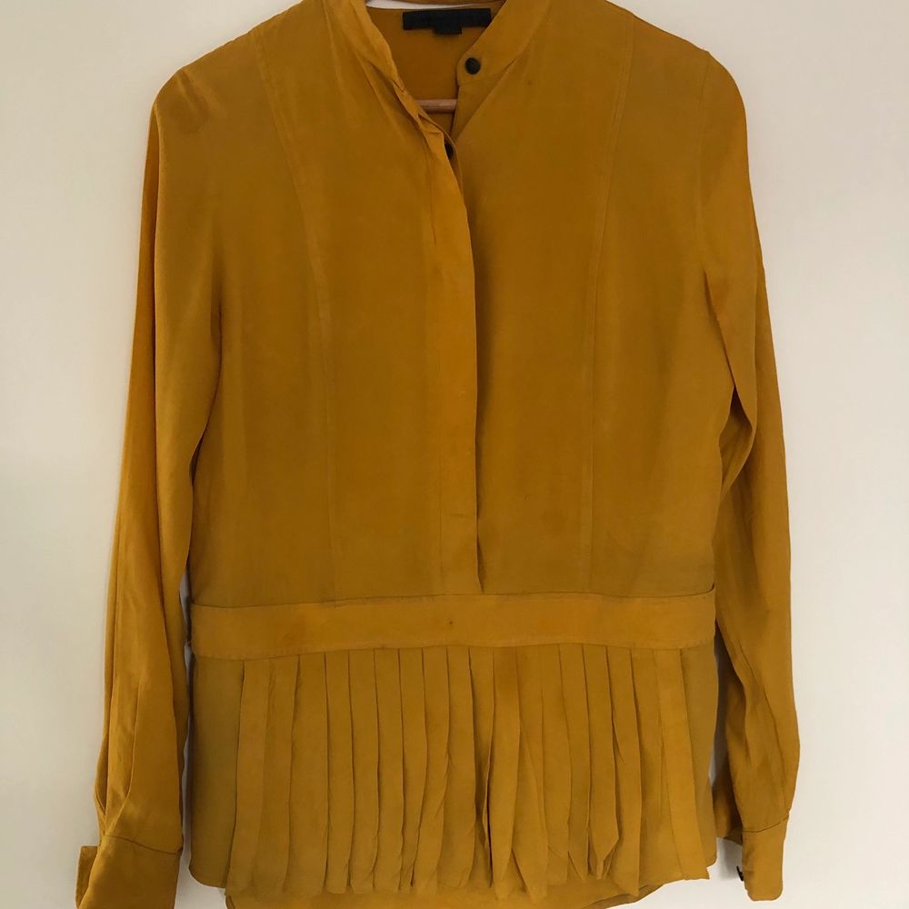 Beautiful blouse in mustard yellow colour. Front features a pleated apron-like detail and hidden button style. Back features split neckline. Fabric shows some signs of aging including decolorization and stains (e.g. around buttons). Skjortor.