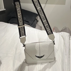 Zadig & Voltaire väska - Unik & snygg äkta zadig väska. Vit glansigt utseende och brett axelremsband. Köpt i London för 370 GP, motsvarar ca 4500kr. Får plats med mycket i. Sparsamt använd, nästan som ny. Kvittot finns inte men äkthetsbevis finns. Köparen står för frakt!