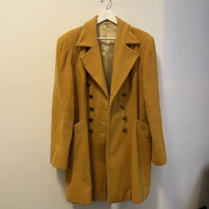 Senapsgul vintagekappa  - En unik kappa från Pura Lana. Dubbelknäppning. Axelpuffar. Senapsgul. Den är inköpt för många många år sen och användes då flitigt så slitage i fodret finns. Vill man se bilder så bara att skicka ett meddelande. Men det synliga på jackan är i fint skick. 