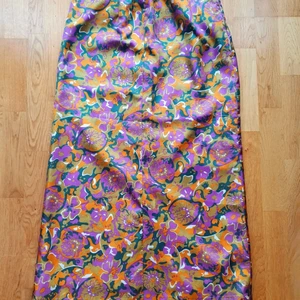 Färgstark långkjol från 70-talet!  - Materialet är mjukt och blankt och antagligen någon typ av konstblandning. Otroligt virvlande blommönster mönster I olivgrönt, mossgrönt, lila, vitt och orange. Vitt satinfoder. Dragkedja I sidan. Superfint skick!  Midja: 42x2 Stuss: 56x2 Längd: 97 cm 