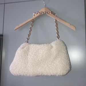 Teddy väska guldfärgad kedja h&m - Supersnygg Teddy väska i beige/vit färg från h&m, ganska stor och rymlig. Aldrig använd . Guldfärgad tjock kedja, väskan har dragkedja 