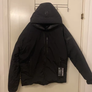 Svart dunjacka från Sail Racing - Snygg svart dunjacka från Sail Racing med huva och dragkedja. Jackan har en stilren design med logotypdetaljer på ärmen och huvan. Perfekt för kyliga dagar. Helt ny jacka aldrig använt skickade in den gamla för reklamation fast fick ett helt ny tillbaks, storlek M 