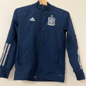 Spansk fotbollsjacka mörkblå - Säljer en mörkblå Spansk träningsjacka från Adidas med dragkedja och långärmade ärmar. Jackan har ett tryckt emblem på bröstet och tre ränder på ärmarna. Perfekt för fotbollsträning eller andra sportaktiviteter. AEROREADY-material för optimal komfort.