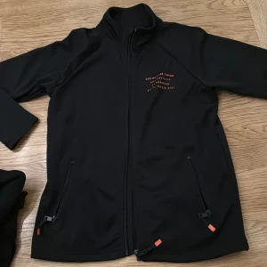 Svart zipkofta från Kappahl - Säljer en svart jacka från Woxo med dragkedja och orange detaljer. Jackan har långa ärmar och två fickor med dragkedjor framtill. Perfekt för en stilren look.