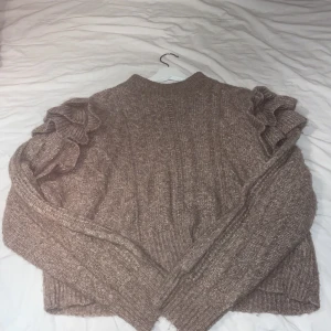 Beige stickad tröja från Gina Tricot - Säljer en mysig beige stickad tröja från Gina Tricot. Tröjan har långa ärmar med volangdetaljer på axlarna och en ribbad struktur. Perfekt för kyliga dagar! Tröjan är över navel så den är lite kort