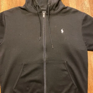 Svart hoodie från Polo Ralph Lauren - Säljer en stilren svart hoodie från Polo Ralph Lauren med dragkedja och det klassiska logotypbroderiet på bröstet. Tröjan har en praktisk huva med dragsko och en stor ficka framtill. Perfekt för en avslappnad look.