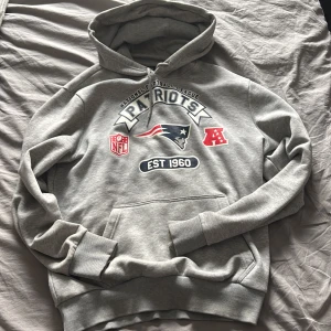 Grå Patriots hoodie från New Era - Säljer en grå hoodie från New Era med Patriots-tryck och NFL-logga. Hoodien har en stor ficka fram och justerbar huva med snören. Perfekt för sportfantasten!