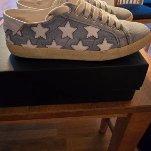 Blå sneakers med stjärnor från Saint Laurent - Snygga blå sneakers från Saint Laurent med vita stjärnor som dekoration. Skorna har en klassisk snörning och är tillverkade i jeansmaterial. Perfekta för en avslappnad stil med en touch av lyx.