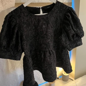 Svart blus från Vero Moda - Snygg svart blus från Vero Moda med puffiga korta ärmar och en unik textur. Perfekt för att ge en elegant touch till din outfit. Blusen har en rund halsringning och en knapp i nacken för enkel påklädning.