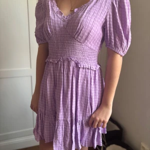 Lila klänning med smock - Säljer en söt lila rutig klänning med v-ringning och korta puffärmar. Klänningen har en smockad midja och volangdetaljer vid fållen. Perfekt för en somrig look! 💜Storlek S 
