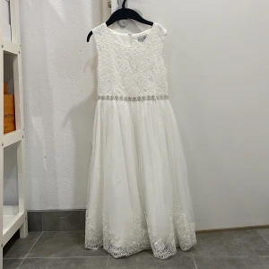 Vit spetsklänning med pärldetaljer - Elegant vit klänning med spetsdetaljer och pärlbroderi i midjan. Klänningen är ärmlös och har en vacker rosett baktill. Kjolen är lång och dekorerad med spets längs nederkanten. Perfekt för speciella tillfällen.