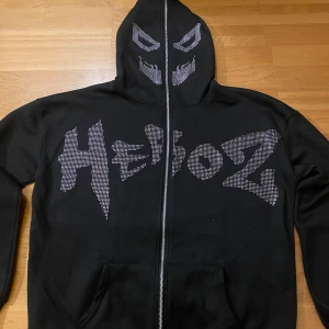 Svart hoodie med dragkedja och vingmönster - Snygg svart hoodie med dragkedja och coolt vingmönster på ryggen. Framsidan har texten 'Heboz' i ett unikt typsnitt. Perfekt för en avslappnad stil med en twist. 