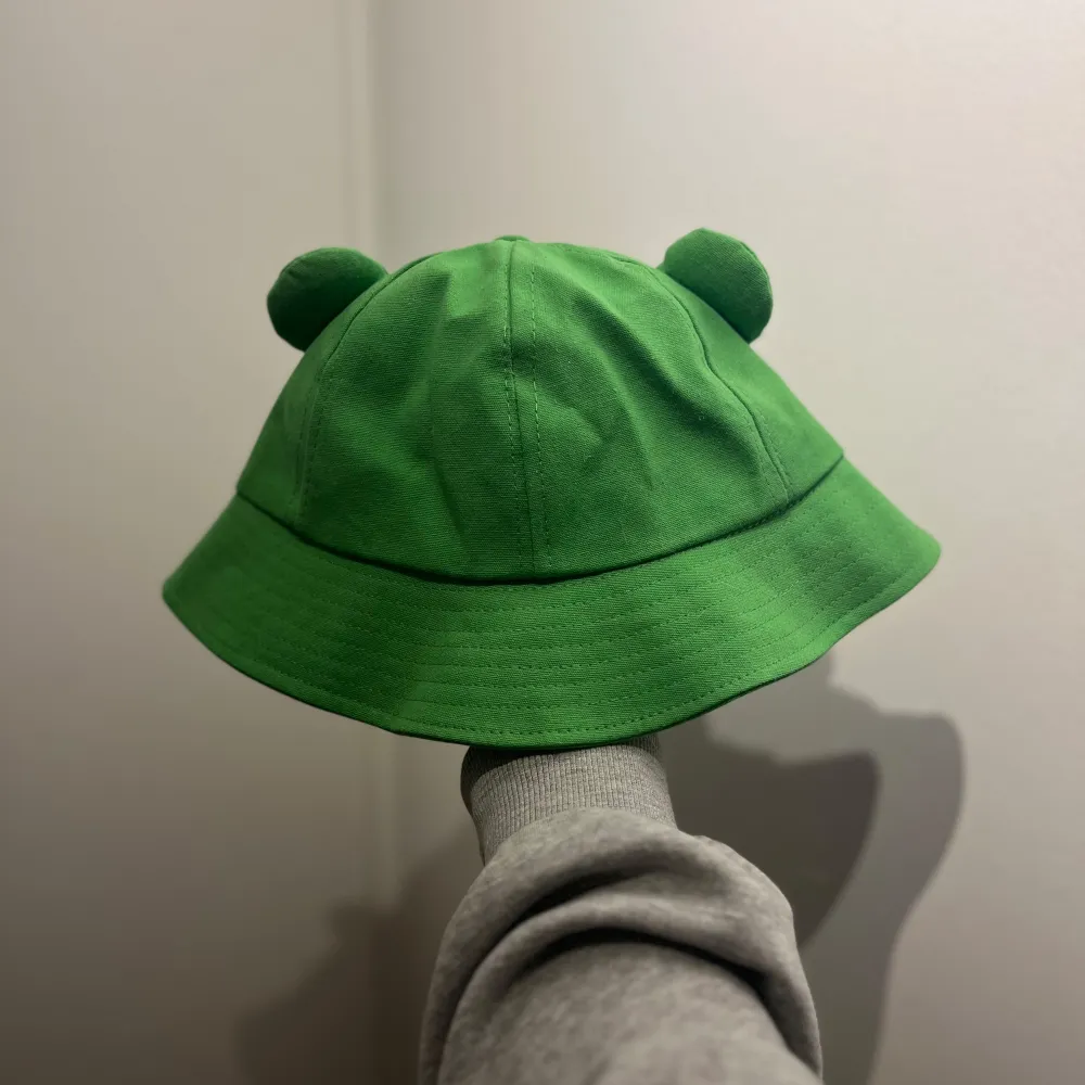 Söt grön bucket hat med grodmotiv och utstickande ögon. Perfekt för en lekfull stil eller som en rolig accessoar. . Asusteet.
