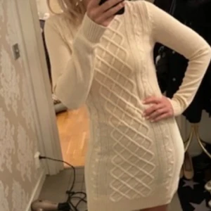 Beige stickad klänning - Säljer en snygg beige stickad klänning med ett flätat mönster framtill. Klänningen är tight och har långa ärmar, perfekt för kyligare dagar. Den är kort och passar bra till både vardag och fest. Ett måste i garderoben för den som älskar stil och komfort!