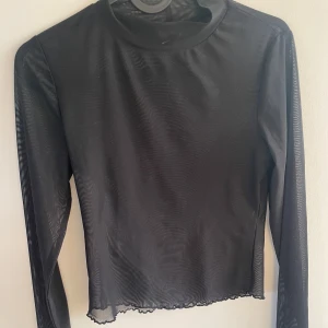 Svart mesh topp från Gina Tricot - Säljer en svart mesh topp från Gina Tricot. Toppen har långa ärmar med subtila volangdetaljer vid fållen. Perfekt för lager-på-lager eller som ett statement-plagg.