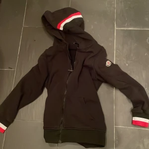 Svart hoodie från Moncler - Säljer en svart hoodie från Moncler med dragkedja och huva. Den har röda och vita detaljer på huvan och ärmsluten. Perfekt för en stilren och bekväm look. Den e ganska använd men väldigt fin 