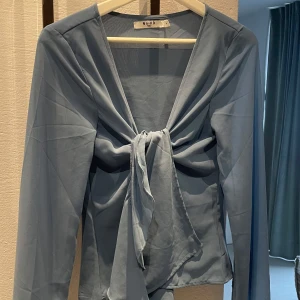 Blå blus från NA-KD - Säljer en söt blå blus från NA-KD med lång ärm och knytning framtill. Sparsamt använd. Blusen är lite skir, alltså lite genomskinlig.
