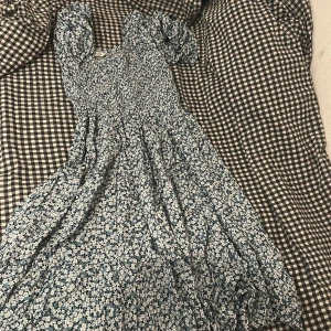Blommig klänning från Pigalle - Säljer en söt blommig klänning från Pigalle i storlek XS. Klänningen har en smockad överdel och korta ärmar. Perfekt för sommarens alla tillfällen med sitt fina blommönster i blått och vitt.