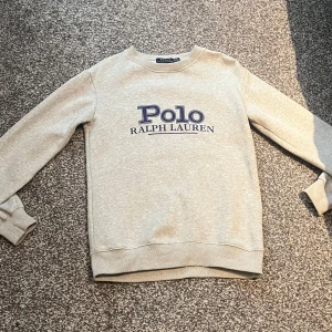 Grå sweatshirt från Ralph Lauren - Säljer en stilren beige sweatshirt från Ralph Lauren med deras klassiska logga i blått på framsidan. Tröjan har långa ärmar och ribbade muddar vid ärmslut och nederkant. Perfekt för en avslappnad stil.
