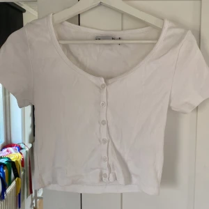 Vit ribbad kortärmad topp med knappar - Säljer en söt vit ribbad topp med korta ärmar och knappar framtill. Perfekt för en avslappnad stil. Toppen har en croppad passform och är superbekväm att bära.