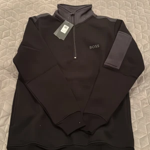 Svart tröja med dragkedja från Boss - Säljer en stilren svart tröja från Boss med dragkedja och hög krage. Tröjan har långa ärmar och en diskret logga på bröstet. Perfekt för en avslappnad men snygg look.