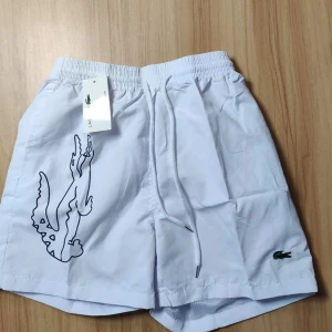 Vita shorts från Lacoste - Snygga vita shorts från Lacoste med elastisk midja och snörning. Stort krokodilmärke på sidan och en bakficka. Perfekta för en avslappnad stil. Kan sänka pris!