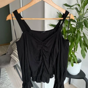 Svart topp med volangdetaljer - Snygg svart topp med breda axelband och volangdetaljer vid fållen. Toppen har en rynkad detalj framtill som ger en fin passform. Perfekt för en stilren look.