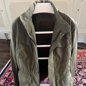 Olivgrön jacka från Massimo Dutti - Säljer en stilren olivgrön jacka från Massimo Dutti. Jackan har dragkedja och knappar framtill, samt praktiska fickor på bröstet. Perfekt för höst och vår med sin lätta design och långa ärmar.