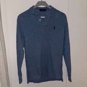 Blå pikétröja från Ralph Lauren - Säljer en stilren blå pikétröja från Ralph Lauren med lång ärm och klassisk krage. Tröjan har knappar vid halsen och den ikoniska loggan broderad på bröstet. Perfekt för en avslappnad men ändå elegant look.