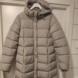 Lång vinterjacka i storlek M - Lång vinterjacka i storlek M. Varm och skön. Fickor på sidorna, dragkedja och knappar fram. Med luva. Helt ny jacka, med prislapp kvar.
