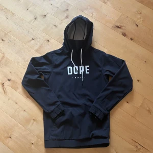 Svart hoodie från Dope - Säljer en svart hoodie från Dope med vit text på bröstet. Storlek XS men passar även S. Tröjan har en justerbar huva med vita snören och en stor ficka framtill. Perfekt för en avslappnad stil.