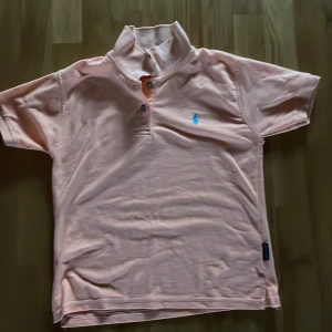 Orange pikétröja från Ralph Lauren - Säljer en snygg rosa pikétröja från Ralph Lauren med klassisk krage och knappdetalj. Tröjan har en broderad logga i blått på bröstet. Perfekt för en stilren look!