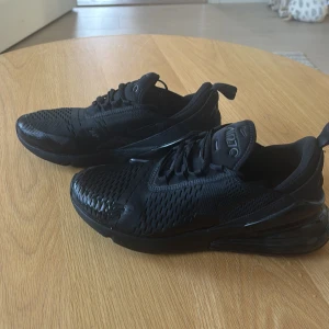 Svarta Nike sneakers 270 - Säljer ett par helsvarta Nike sneakers med snörning och en modern design. Skorna har en luftig ovandel och en synlig luftkudde i hälen för extra komfort. Perfekta för både träning och vardag. Bara använda 1 gång