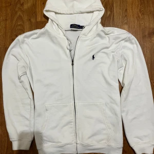 Vit hoodie från Ralph Lauren - Säljer en stilren vit hoodie från Ralph Lauren med dragkedja och broderad logga på bröstet. Perfekt för en avslappnad look.  Hoodien är i top skick 9/10. För mera information hör gärna av er ✅✅