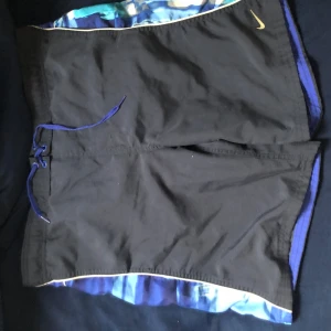 Svarta och blå badbyxor från Nike - Snygga svarta badbyxor från Nike med blå detaljer och ett coolt mönster på sidorna. De har en praktisk snörning i midjan för bästa passform. Perfekta för sommarens alla äventyr!