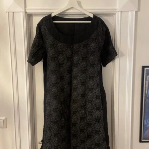 Svart klänning med blommigt mönster - Snygg svart klänning med ett subtilt blommigt mönster. Den har korta ärmar och en rund halsringning. Perfekt för en stilren look. Från UNO, storlek L, två fickor vid sidan. 