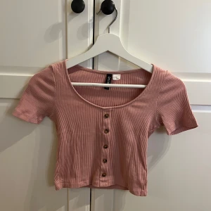 Rosa ribbad topp från H&M Divided - Säljer en söt rosa ribbad topp från H&M Divided. Toppen är croppad med korta ärmar och har knappar framtill. Perfekt för en avslappnad stil.