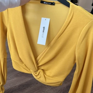 Gul omlottopp från Gina Tricot - Snygg gul omlottopp från Gina Tricot med långa ärmar och knytdetalj framtill. Perfekt för att liva upp garderoben med en färgklick. Toppen har en v-ringad design som ger en stilren look. Kunden står för frakten/ alternativt mötas upp i Norrköping!