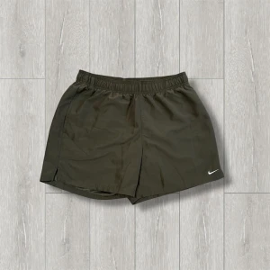 Olivgröna träningsshorts från Nike - Säljer ett par olivgröna träningsshorts från Nike. De har en elastisk midja och är tillverkade i polyester för en bekväm passform. Perfekta för träning eller en aktiv dag. Nike-loggan syns diskret på benet.