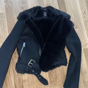 Svart jacka med pälsdetaljer - Snygg svart jacka från River Island med pälsdetaljer på kragen och insidan. Jackan har dragkedjor och ett bälte i midjan för en cool look. Perfekt för kyligare dagar. Storlek S