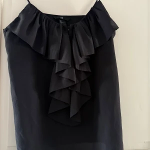 Svart blus från H&M - Snygg svart blus från H&M med volangdetaljer framtill. Jättesöt på och jag säljer den då den inte kommer till användning längre 
