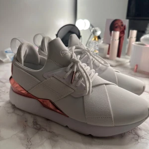 Vita sneakers med roségulddetaljer från Puma - Snygga vita sneakers från Puma med roségulddetaljer på hälen och snörning. Skorna har en modern design med elastiska band över ovandelen och en bekväm sula. Perfekta för en stilren look. Sköna skor att träna eller promenera i. Har dock inte kommit till användning så mycket. 