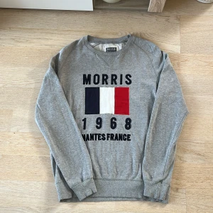Grå sweatshirt från Morris - Säljer en grå sweatshirt från Morris med tryck av franska flaggan och texten 'Morris 1968 Nantes France'. Tröjan har långa ärmar och ribbade muddar vid ärmslut och nederkant. Perfekt för en avslappnad stil.