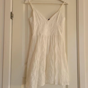 Vit klänning med broderad spets - Charmig vit klänning med v-ringad design och smala axelband. Klänningen har ett vackert broderat spetsmönster och en dragkedja i ryggen. Perfekt för sommarens alla tillfällen!